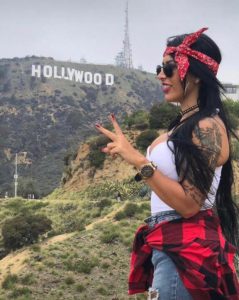 foto de moça com bandana na cabeça fazendo o sinal da paz com as maos e o letreiro de hollywood ao fundo